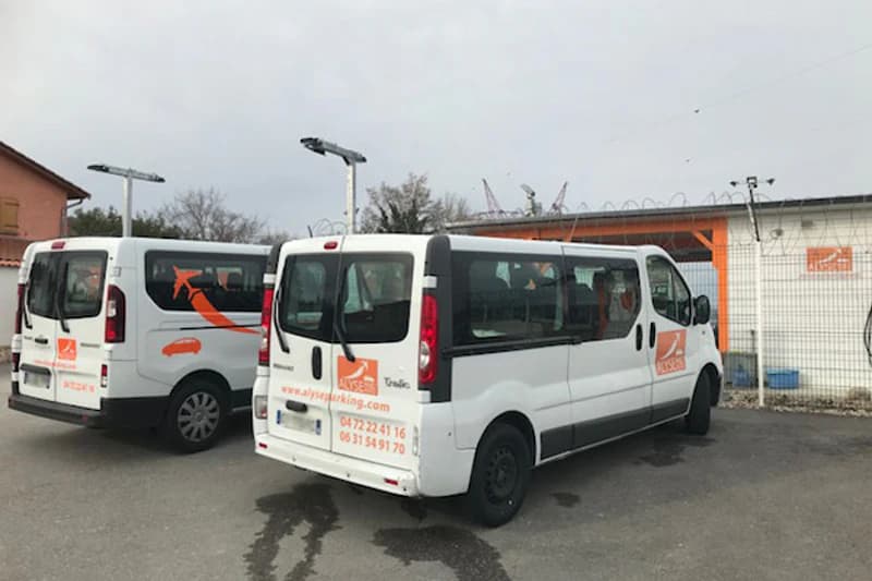 Zwei weiße Shuttlebusse mit Alyse-Logo auf eingezäuntem Parkplatz vor Lagergebäude.
