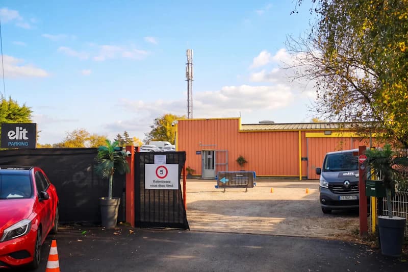 Einfahrt zu umzäuntem Parkplatz mit offenem Tor, Tempo-5-Schild, oranger Halle und geparkten Fahrzeugen.