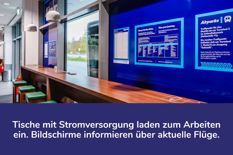 Langer Holztisch mit Hockern vor Fenstern und blauen Informationsbildschirmen an der Wand.