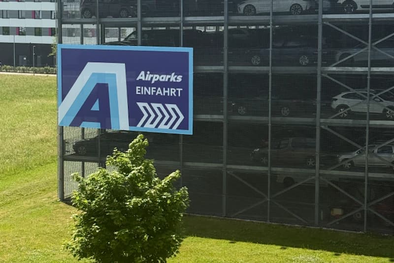 Mehrstöckiges Parkhaus mit großem Schild Airparks Einfahrt und Pfeilen, im Vordergrund Rasen und Baum.