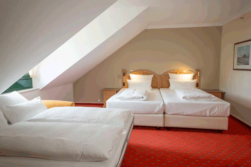 Dachzimmer mit drei Einzelbetten, rotem Teppich, schrägem Fenster und zwei Nachtkästchen.