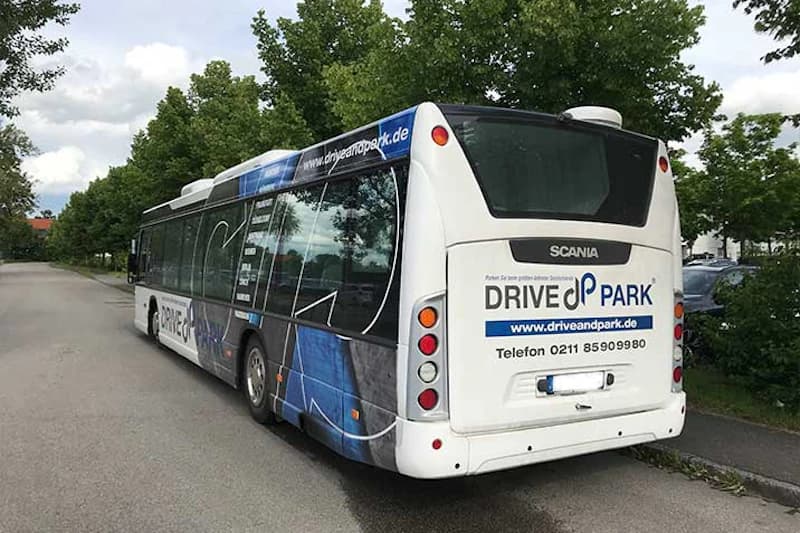 Weißer Shuttlebus mit drive&park Werbung hinten und seitlich, Scania-Logo, am Straßenrand neben Bäumen geparkt.