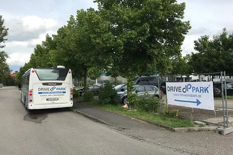 Bus mit DRIVE&PARK-Logo neben Zaun mit Banner und Pfeil, dahinter geparkte Autos unter Bäumen.