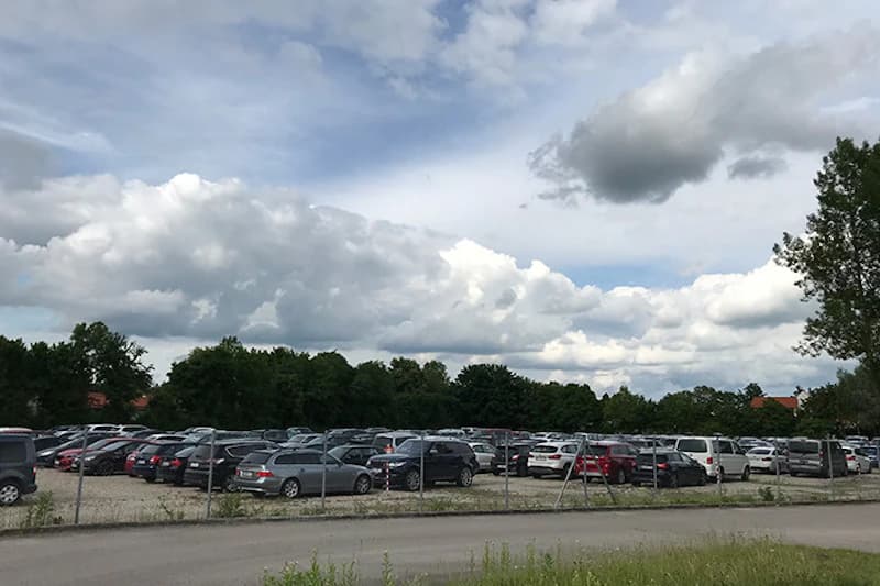 Gezaeunter Schotterparkplatz mit zahlreichen abgestellten Autos und Baumreihe unter bewölktem Himmel.