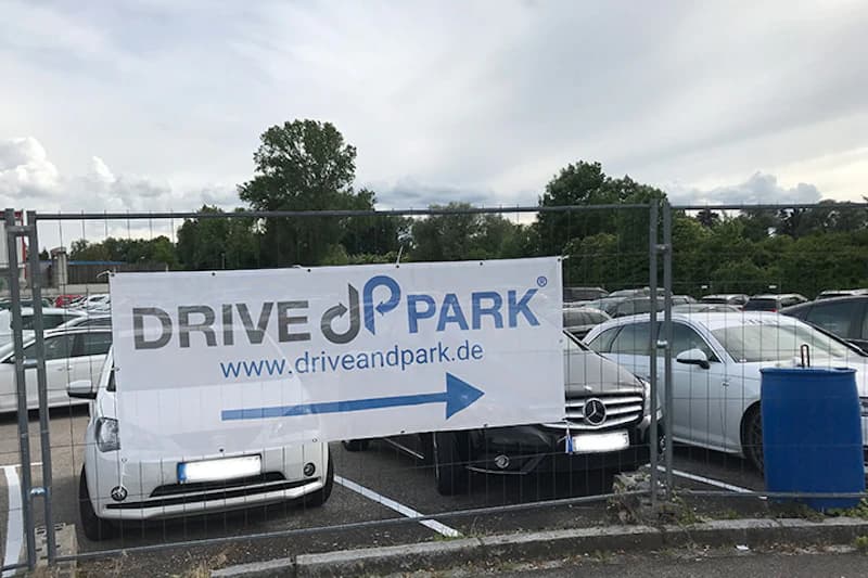 Metallzaun mit Drive&Park-Banner und Richtungspfeil vor geparkten Autos auf einem Parkplatz.