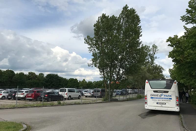 Straße und Gehweg neben eingezäuntem Parkplatz mit zahlreichen Autos und weißem Shuttlebus von Drive&Park.