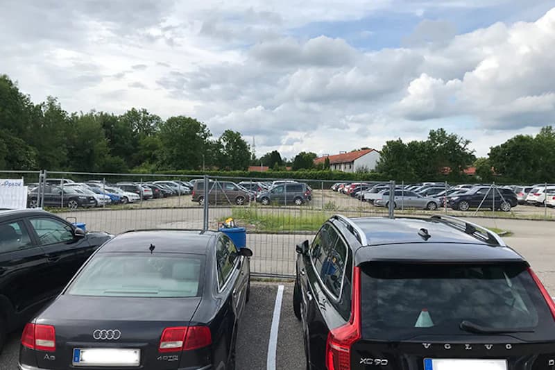 Eingezäunter Parkplatz mit mehreren geparkten Autos, Metallzaun im Vordergrund und Bäume unter bewölktem Himmel.