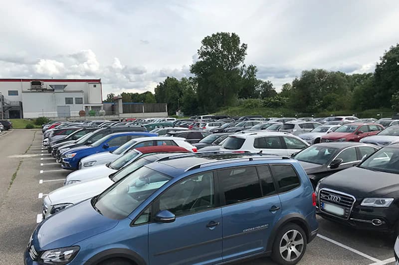 Großer Außenparkplatz mit dicht geparkten Autos, markierten Stellplätzen, Gebäude links und Bäumen unter bewölktem Himmel.