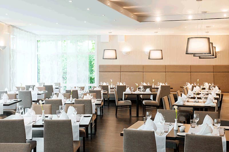 Helles Hotelrestaurant mit gedeckten Tischen, grauen Stühlen, Tischwäsche und großen Fenstern.