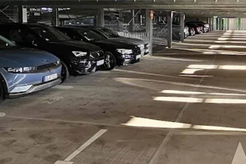 Überdachtes mehrgeschossiges Parkhaus mit links geparkten Autos, Betonstützen und Lichtflecken auf dem Boden.