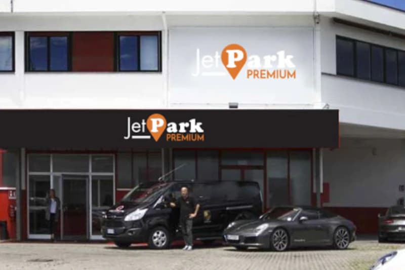 Fassade mit JetPark Premium Schild, schwarzer Transferbus, stehender Mitarbeiter und grauer Sportwagen vor dem Eingangsbereich.