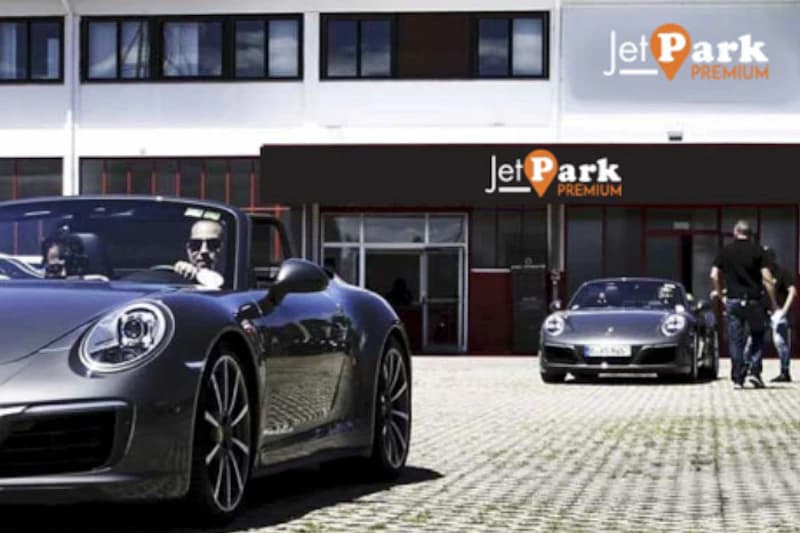 Zwei graue Sportwagen vor dem Eingang eines JetPark Premium Parkplatzgebäudes, Personen stehen daneben.