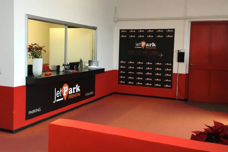 Empfangstresen JetPark Premium mit Schalterfenster, Computer, Blumenvase und Logowand in rot-weißer Halle.