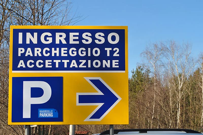 Gelbes Schild mit Aufschrift INGRESSO PARCHEGGIO T2 ACCETTAZIONE, blaues Parksymbol und Pfeil nach rechts vor Bäumen.
