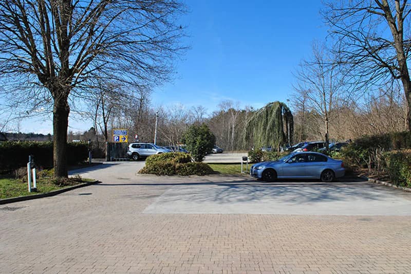 Parkfläche mit einzelnen Autos, bepflanzten Inseln, Bäumen und Parkhinweisschild unter blauem Himmel.