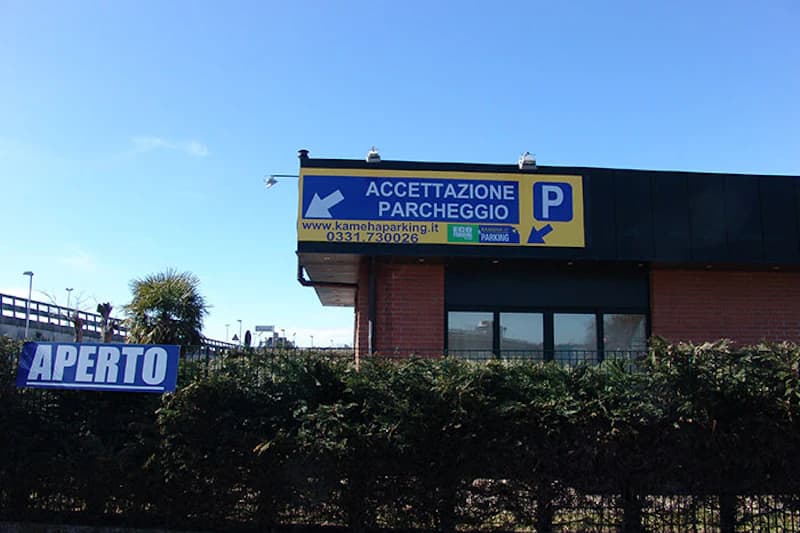 Kleines Backsteingebäude mit blau-gelbem Schild ACCETTAZIONE PARCHEGGIO, APERTO-Banner und Hecke vorne.