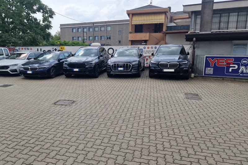 Mehrere geparkte schwarze und dunkelblaue Pkw auf gepflastertem Parkplatz vor Werbezaun und Wohngebäuden.
