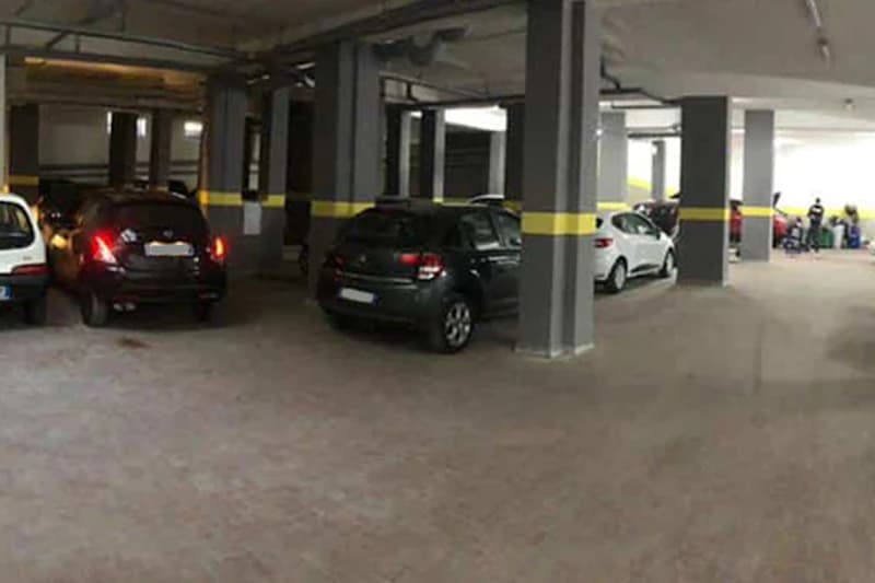 Überdachte Parkhalle mit mehreren geparkten Pkw, graue Säulen mit gelbem Streifen und Personen am Rand.