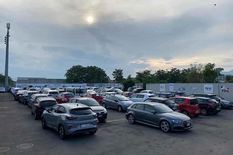 Außenparkplatz mit vielen geparkten Autos, Beleuchtungsmast, umzäunter Perimeter und bewölkter Himmel.