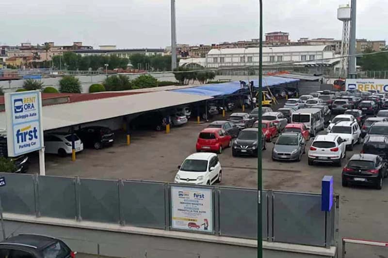 Eingezäunter Parkplatz mit vielen geparkten Autos und überdachten Stellplätzen, First-Schild sichtbar.