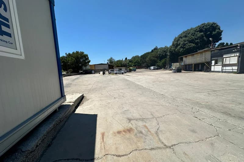 Große leere Betonfläche eines Parkplatzes mit Containern am Rand, einigen Autos in der Ferne und Bäumen im Hintergrund.