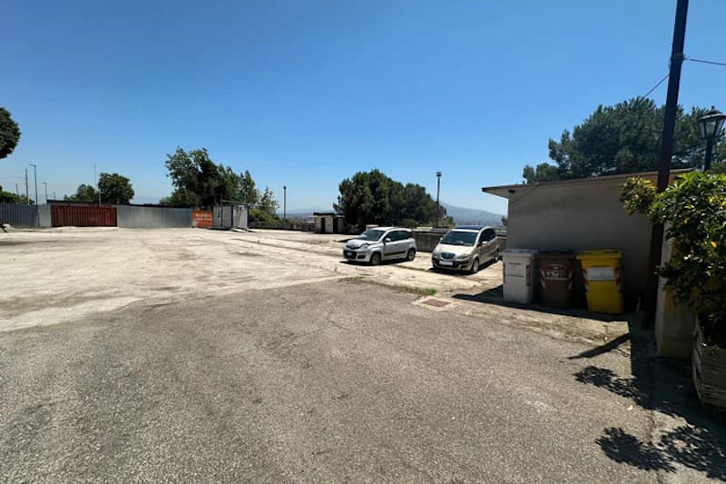 Großer offener Parkplatz mit zwei geparkten Autos, Mülltonnen neben kleinem Gebäude und Bäumen am Rand.
