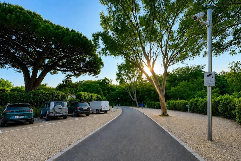 Asphaltweg zwischen Kiesstellplaetzen mit geparkten Autos, Bueschen und Ueberwachungskamera an einem Pfosten.