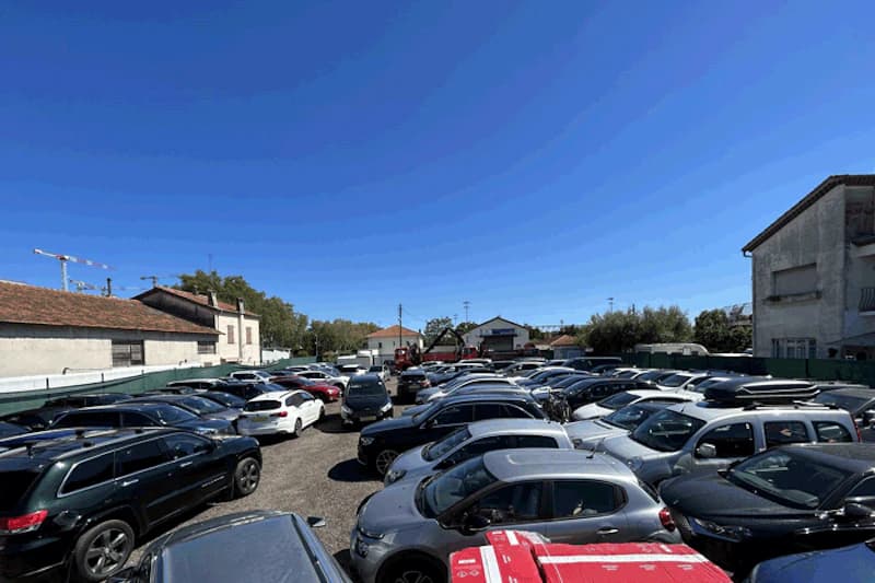 Voller Freiluftparkplatz mit dicht aneinandergereihten Autos, umzäunt und von Gebäuden umgeben unter blauem Himmel.