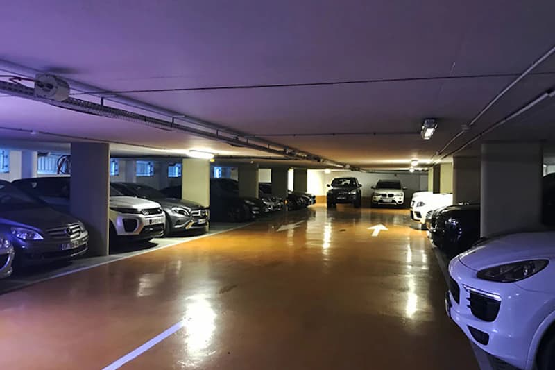 Tiefgaragenebene mit Reihen geparkter Autos zwischen Betonpfeilern und beleuchtetem Gang mit Richtungspfeil auf dem Boden.