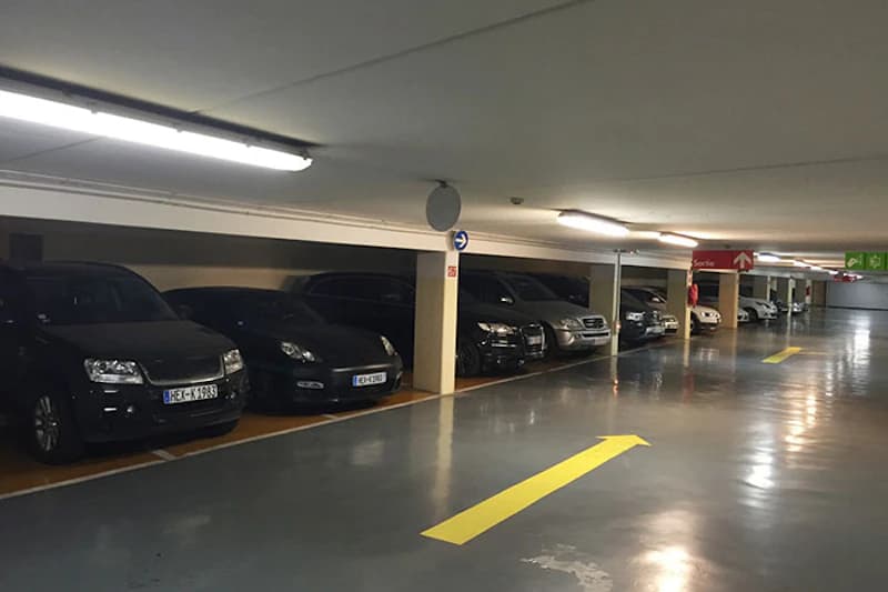 Parkebene in einem Parkhaus mit geparkten Autos, Stützpfeilern, Deckenbeleuchtung und gelbem Fahrpfeil auf dem Boden.