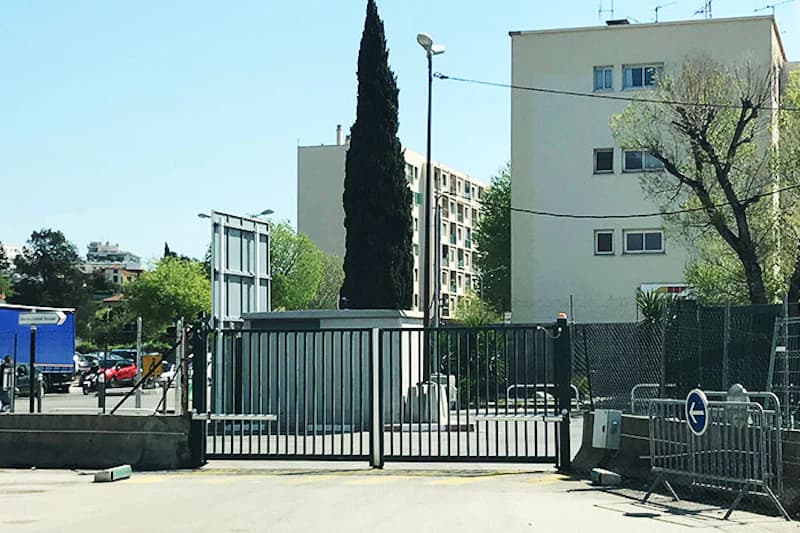 Geschlossenes Metallschiebetor vor umzäuntem Parkplatz, Pförtnerhäuschen, hohe Zypresse und Wohngebäude im Hintergrund.
