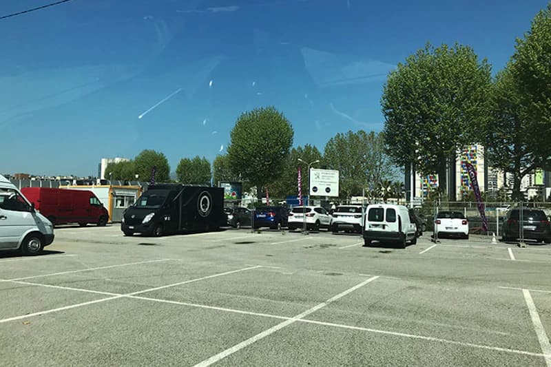 Außenparkplatz mit leeren und belegten Stellflächen, mehreren Transportern und geparkten Autos vor Baumreihe.