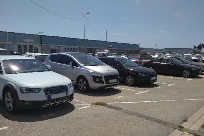Mehrere geparkte Autos auf sonnigem Außenparkplatz vor niedrigen Hallengebäuden.