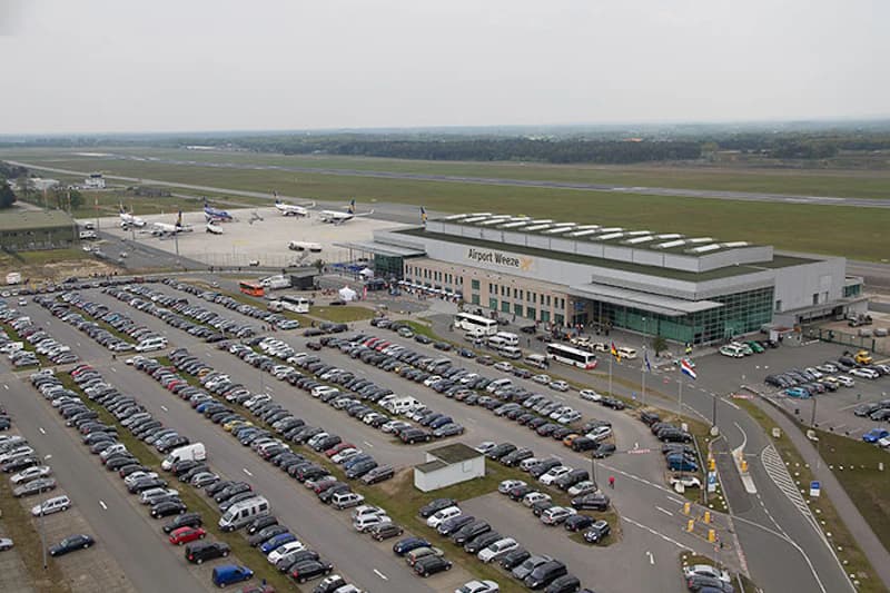 Großer Parkplatz vor Terminal Airport Weeze mit Reihen geparkter Autos, Bussen, Terminalgebäude und Rollfeld.