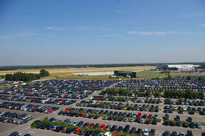 Großer Außenparkplatz mit dicht geparkten Autos und Fahrspuren, im Hintergrund Rollfeld und Terminalgebäude.