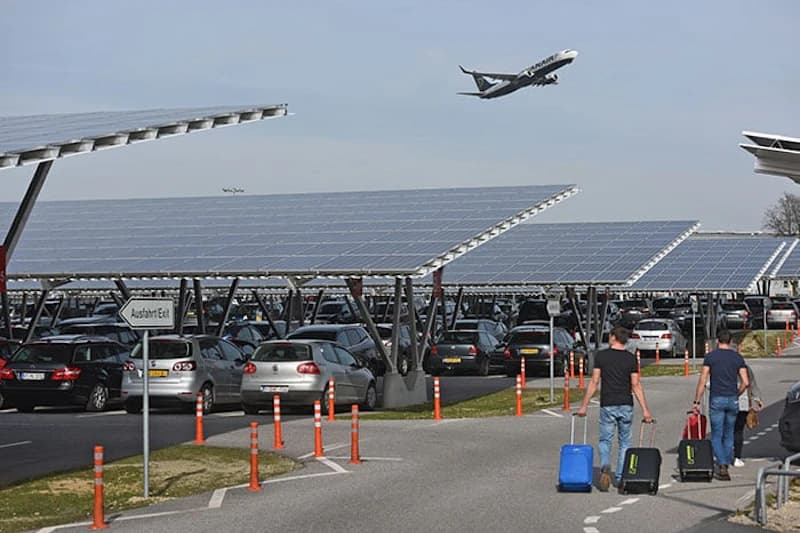 Überdachter Parkplatz mit Solarpaneelen, geparkte Autos, zwei Männer mit Rollkoffern und startendes Flugzeug am Himmel.