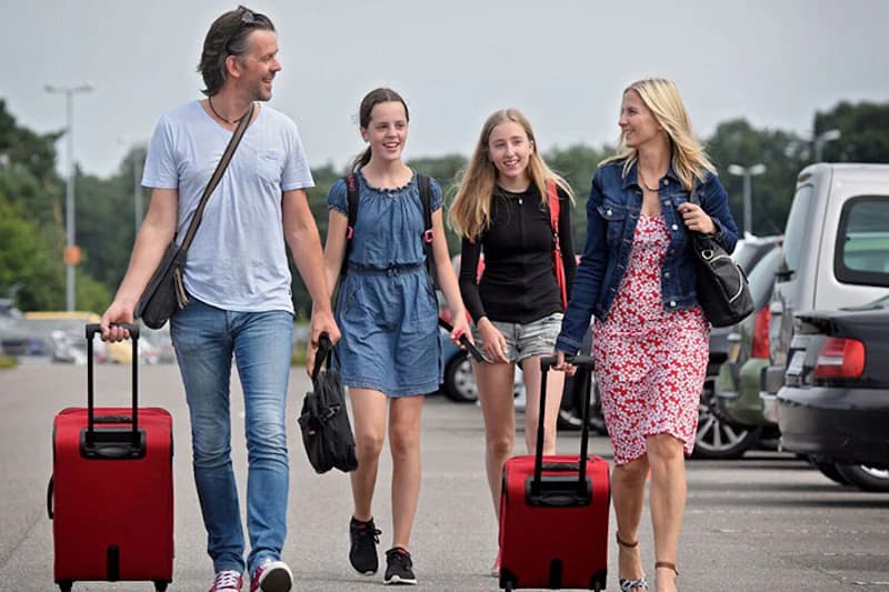 Familie mit zwei Töchtern zieht rote Rollkoffer auf einem Parkplatz neben geparkten Autos.