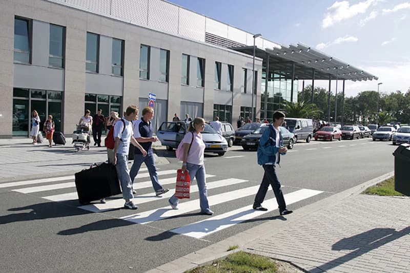 Passanten mit Gepäck überqueren Zebrastreifen vor Terminal mit Ankunftsbereich und geparkten Autos.