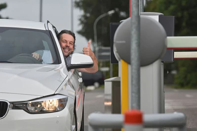 Mann in weißem Auto an Einfahrtsschranke zeigt Daumen hoch.