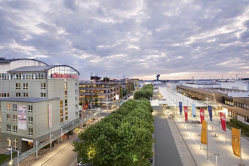 Mövenpick Hotel am Flughafen Nürnberg links, baumgesäumte Zufahrtsstraße und Terminal mit Fahnen unter bewölktem Himmel.
