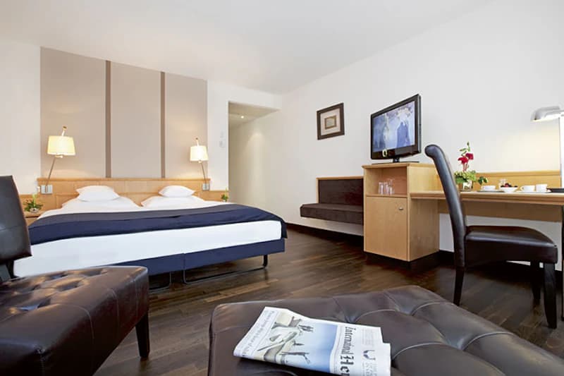 Hotelzimmer mit Doppelbett, Nachttischlampen, Schreibtisch mit Stuhl, Fernseher und Lederhocker mit Zeitung.