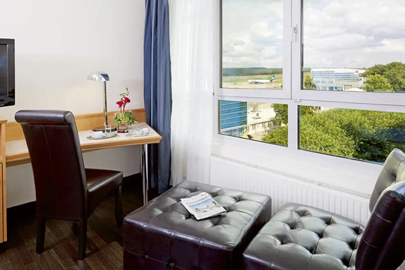 Hotelzimmer mit Schreibtisch, Lederhockern vor grosser Fensterfront und Blick auf Flughafen und Terminal.