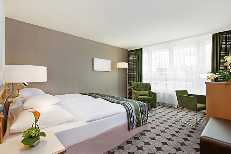 Helles Hotelzimmer mit Doppelbett, zwei Nachttischen, grünem Sessel und Schreibtisch vor großem Fenster.