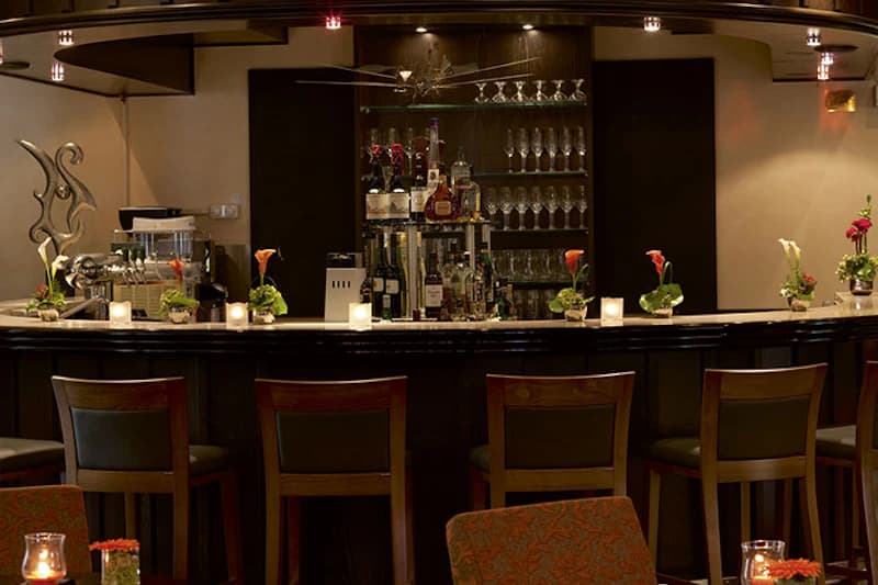 Dunkle Hotelbar mit Tresen, fünf Barhockern, Regal mit Gläsern und Flaschen sowie Kerzen und Blumen.