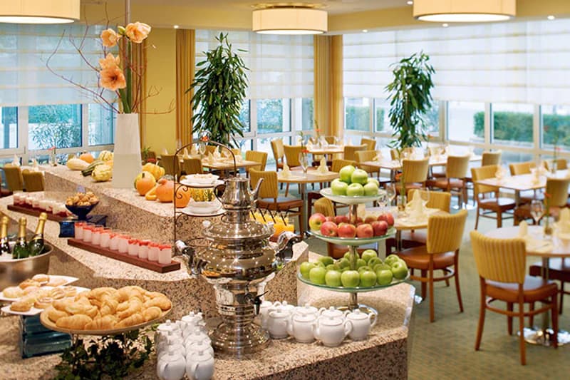 Frühstücksbuffet mit Obst, Gebäck, Teekannen und gedeckten Tischen im Hotelrestaurant.
