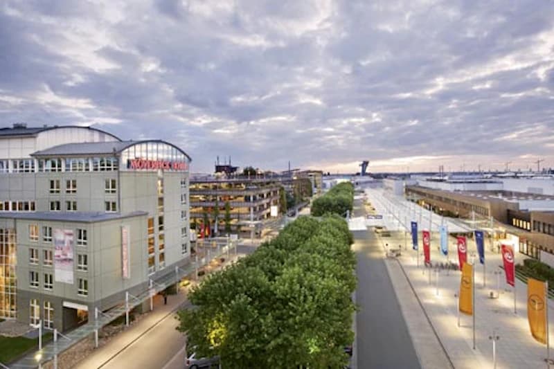 Mövenpick Hotel am linken Bildrand, Baumreihe, Straße und Flughafen mit Fahnen und Terminal unter bewölktem Himmel.