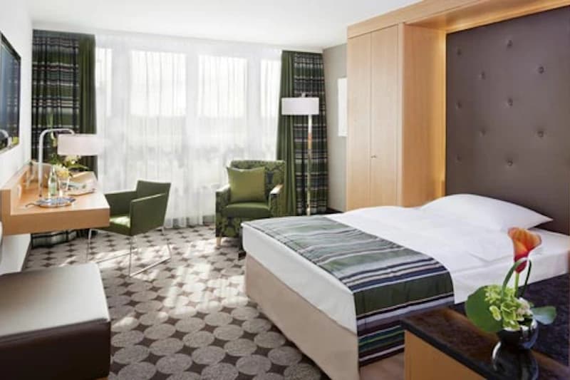 Helles Hotelzimmer mit Doppelbett, gepolstertem Kopfteil, Fenster mit Gardinen, zwei Sesseln, Schreibtisch und Blumen.