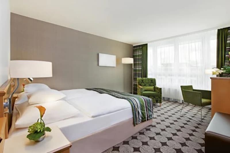 Hotelzimmer mit Doppelbett, zwei Nachttischen, Leselampen, grünem Sessel, Schreibtisch und großem Fenster mit Gardinen.