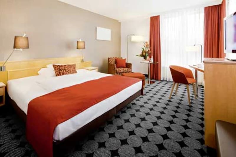 Großes Hotelzimmer mit Doppelbett, rotem Bettläufer, Sessel mit Hocker, Schreibtisch und großem Fenster.