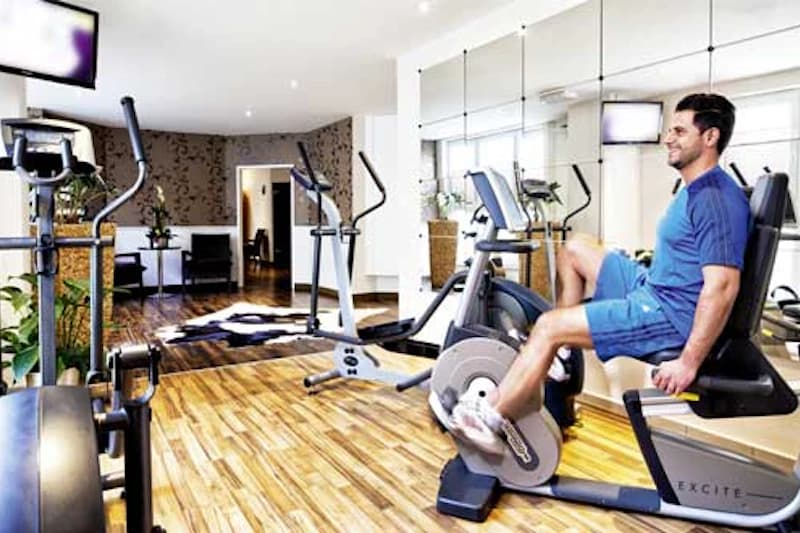 Mann trainiert auf Liegefahrrad in hellem Fitnessraum mit Spiegelwand und weiteren Trainingsgeräten.